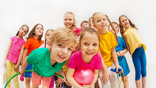 Das Bild zeigt 10 Kinder, die alle in die Kamera schauen. Viele von Ihnen haben Kleingeräte, wie z.B. Springseil, Gymnastikball oder Reifen in der Hand.