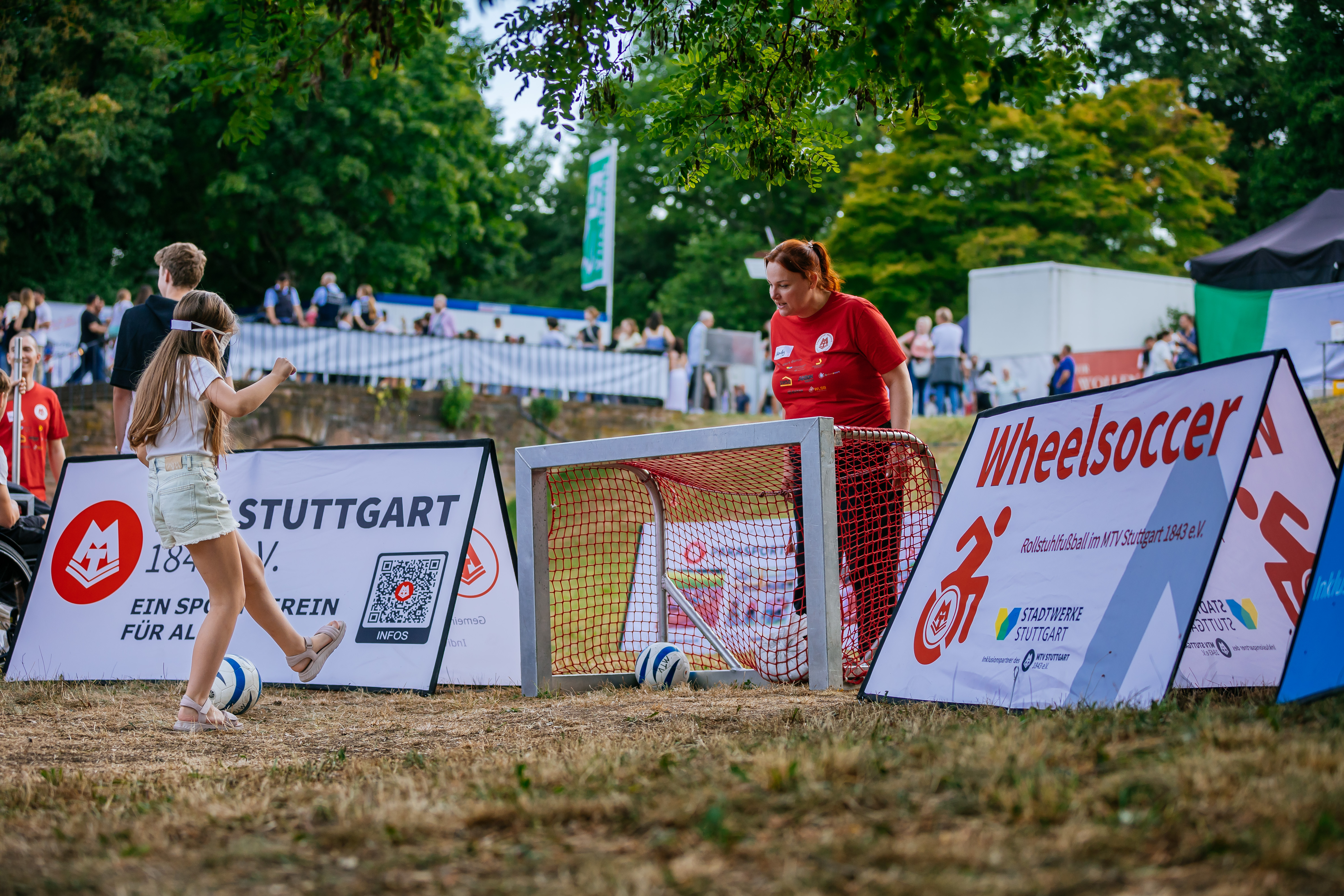 Kind mit verbundenen Augen versucht sich im Blindenfußball. Bewegt sich vom Betrachter weg auf ein kleines Kindertor hinter dem eine Frau steht und Anweisungen gibt.