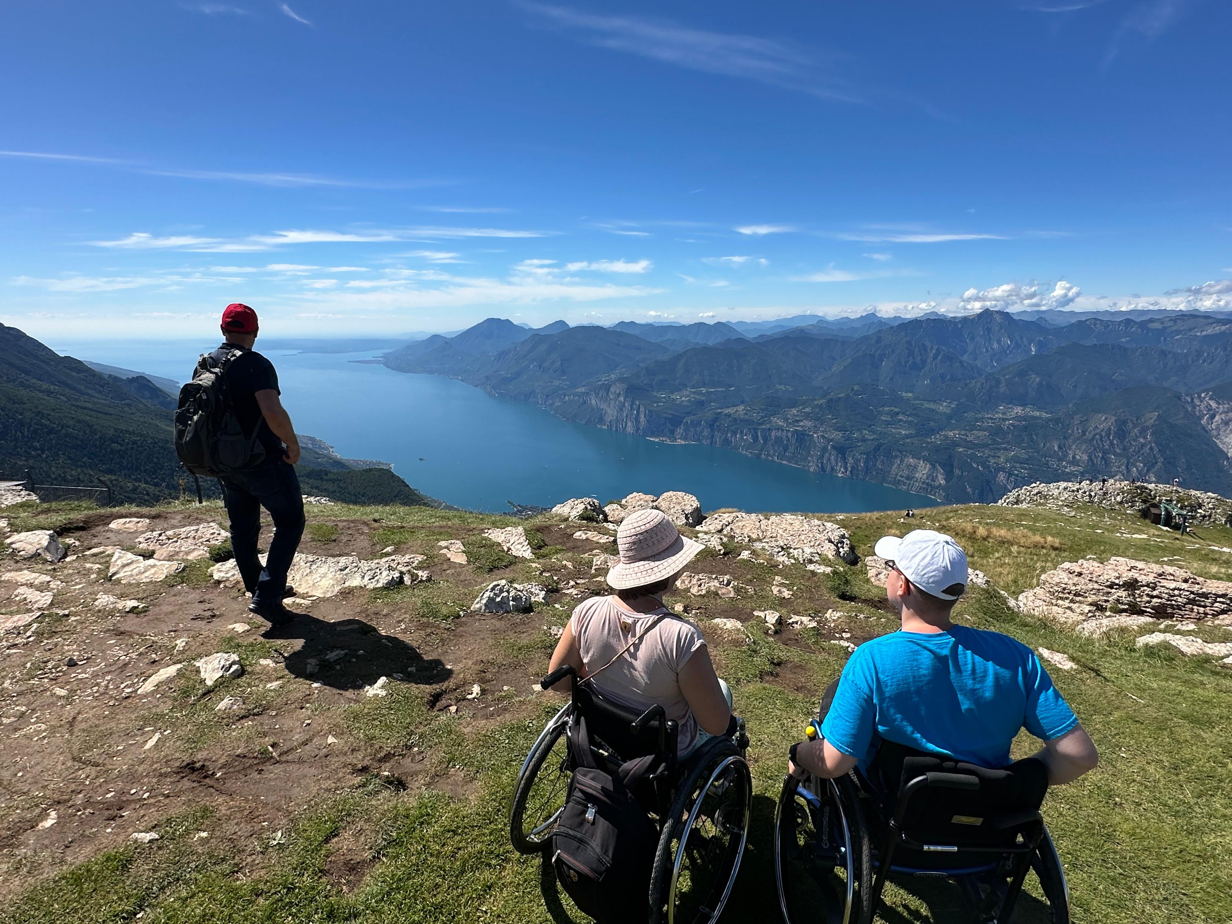 Grandiose Aussicht vom Monte Baldo auf den Gardasee. Zwei Rollstuhlnutzende sehen hinab
