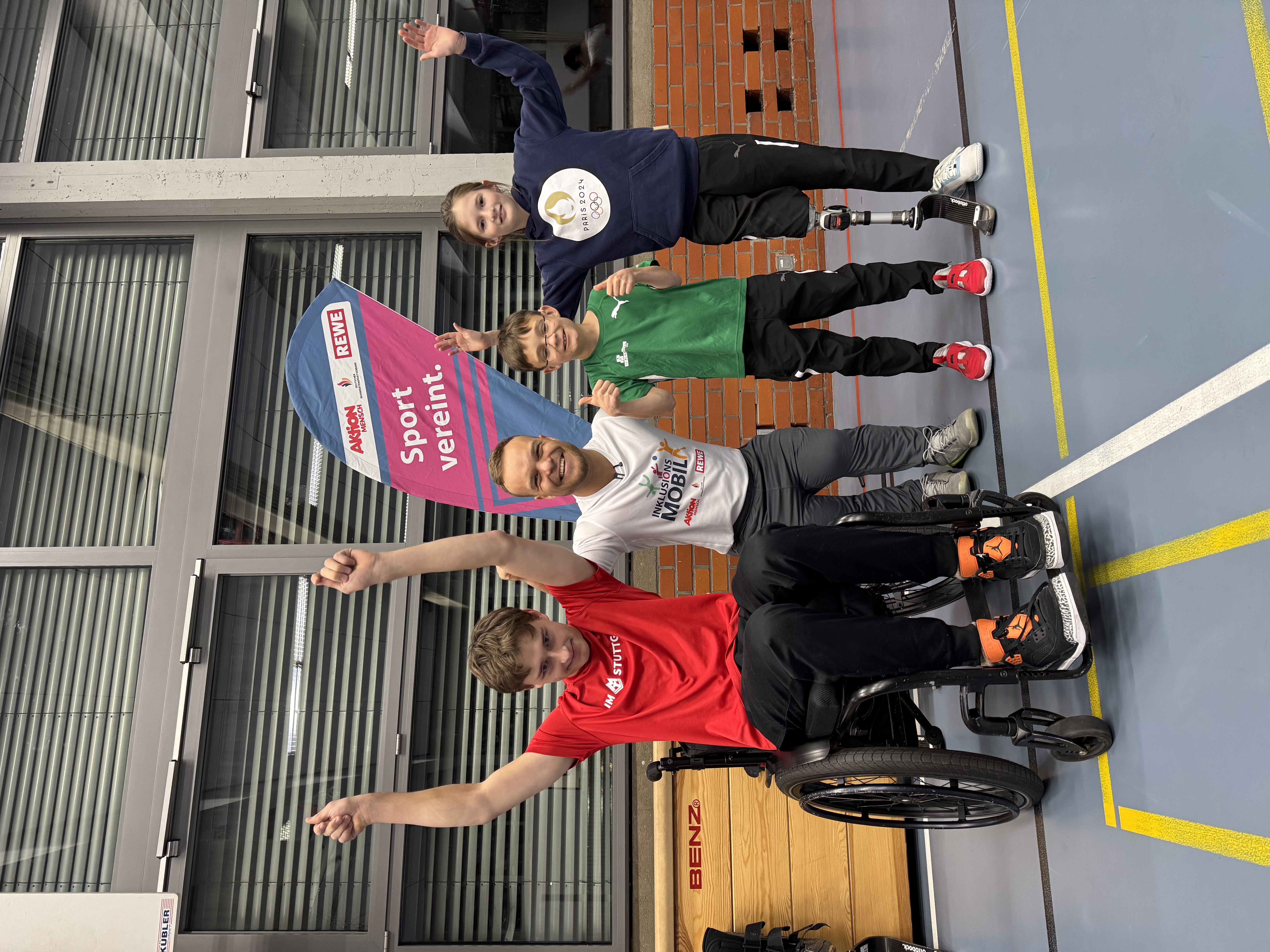Vier Sportler*innen - ein jugendlicher Rollstuhlfahrer, Niko Kappel, ein Kind und ein Kind mit Oberschenkelprothese rechts - jubeln vor einer Turnhallenfensterwand und vor einer Beachflag der Aktion Mensch, des Deutschen Behindertensportverbands und REWE mit dem Titel "Sport vereint". 