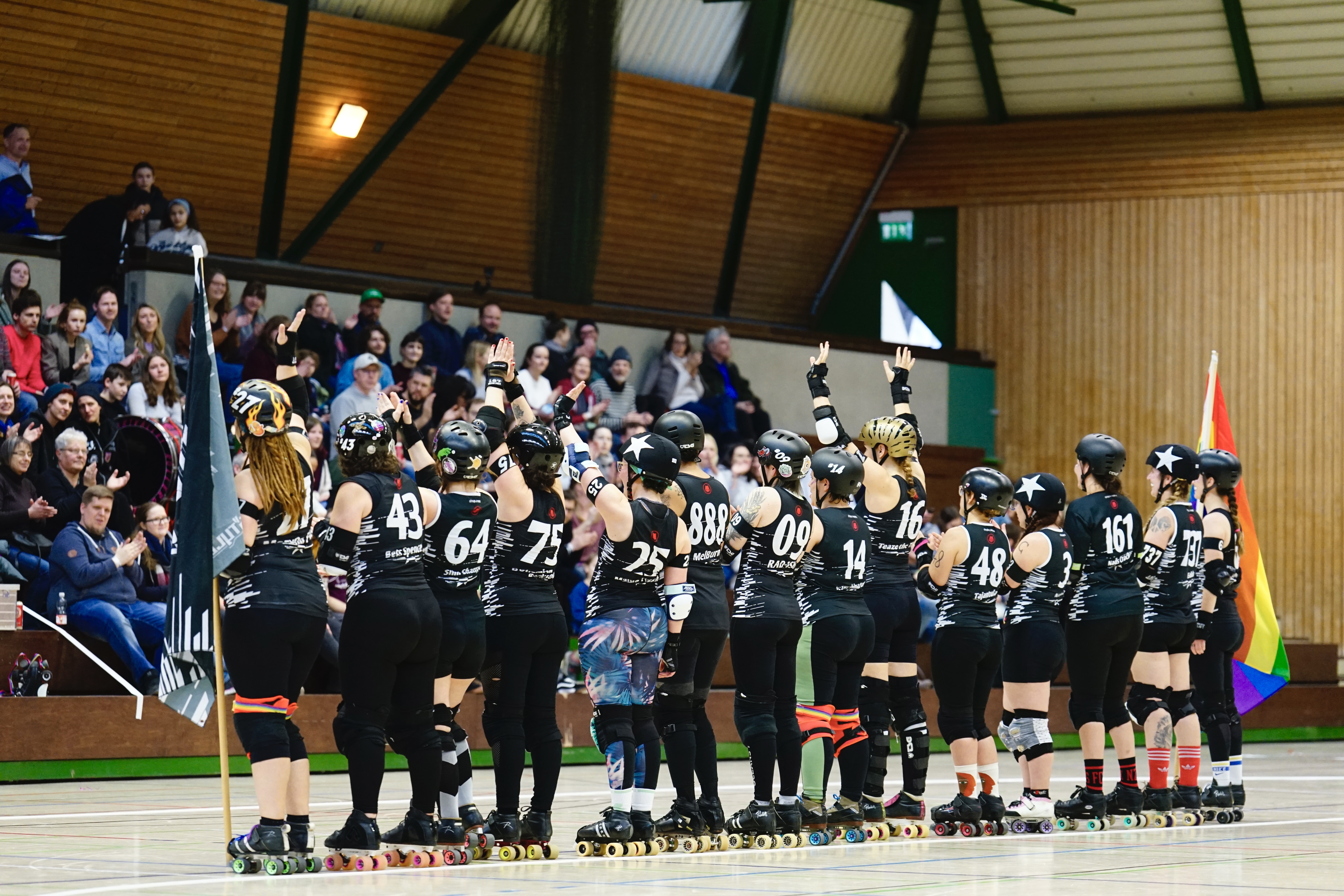 Das Bild zeigt Roller-Derby-Spielerinnen, die vor dem Spiel aufgereiht nebeneinanderstehen. Sie blicken zum Publikum und lassen sich beklatschen.