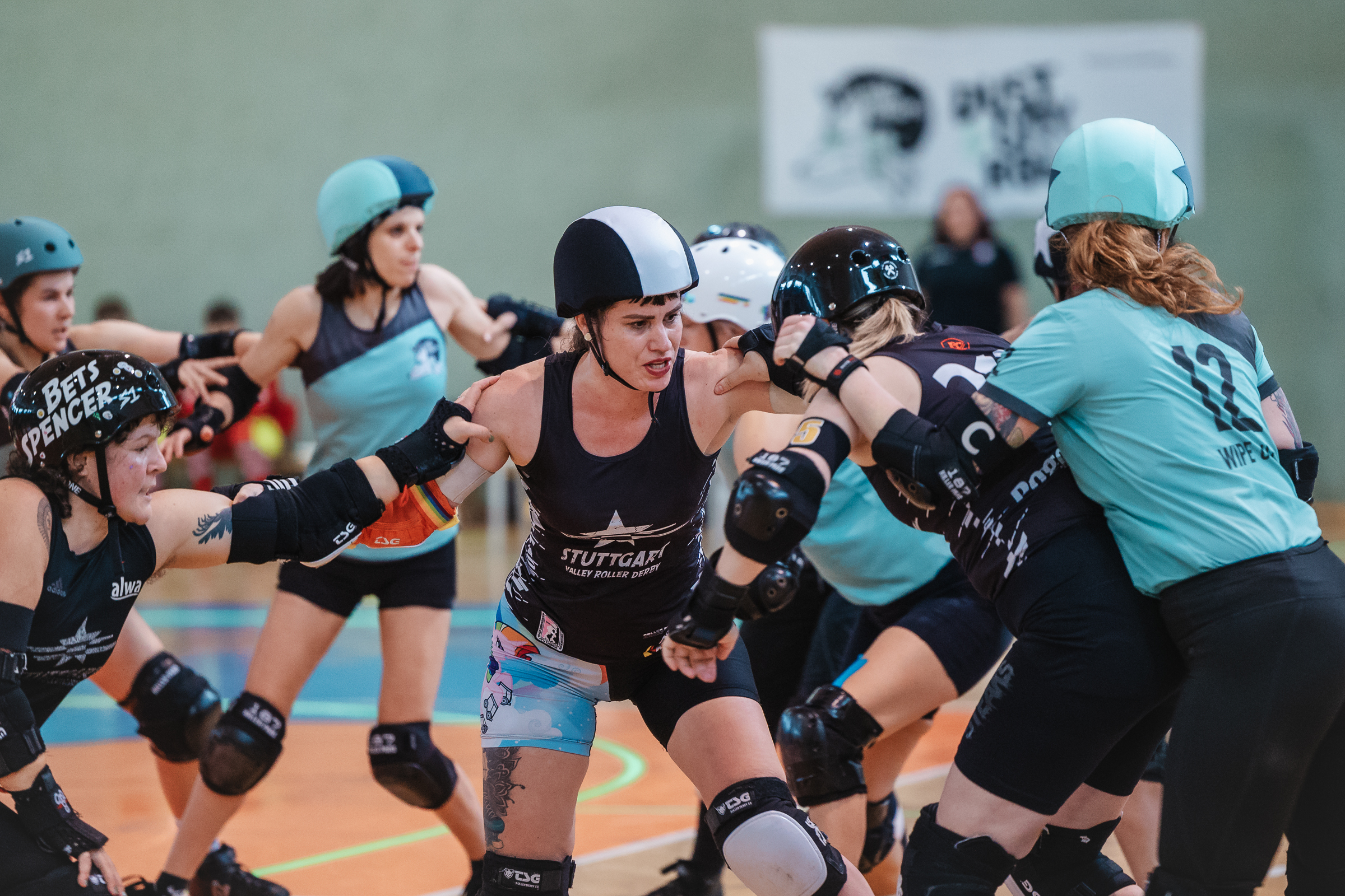Das Bild zeigt ein Roller-Derby-Team, das gerade die Gegnerin abwehrt. Die Spielerinnen halten sich aneinander fest und man sieht die Kraftanstrengung im Gesicht.