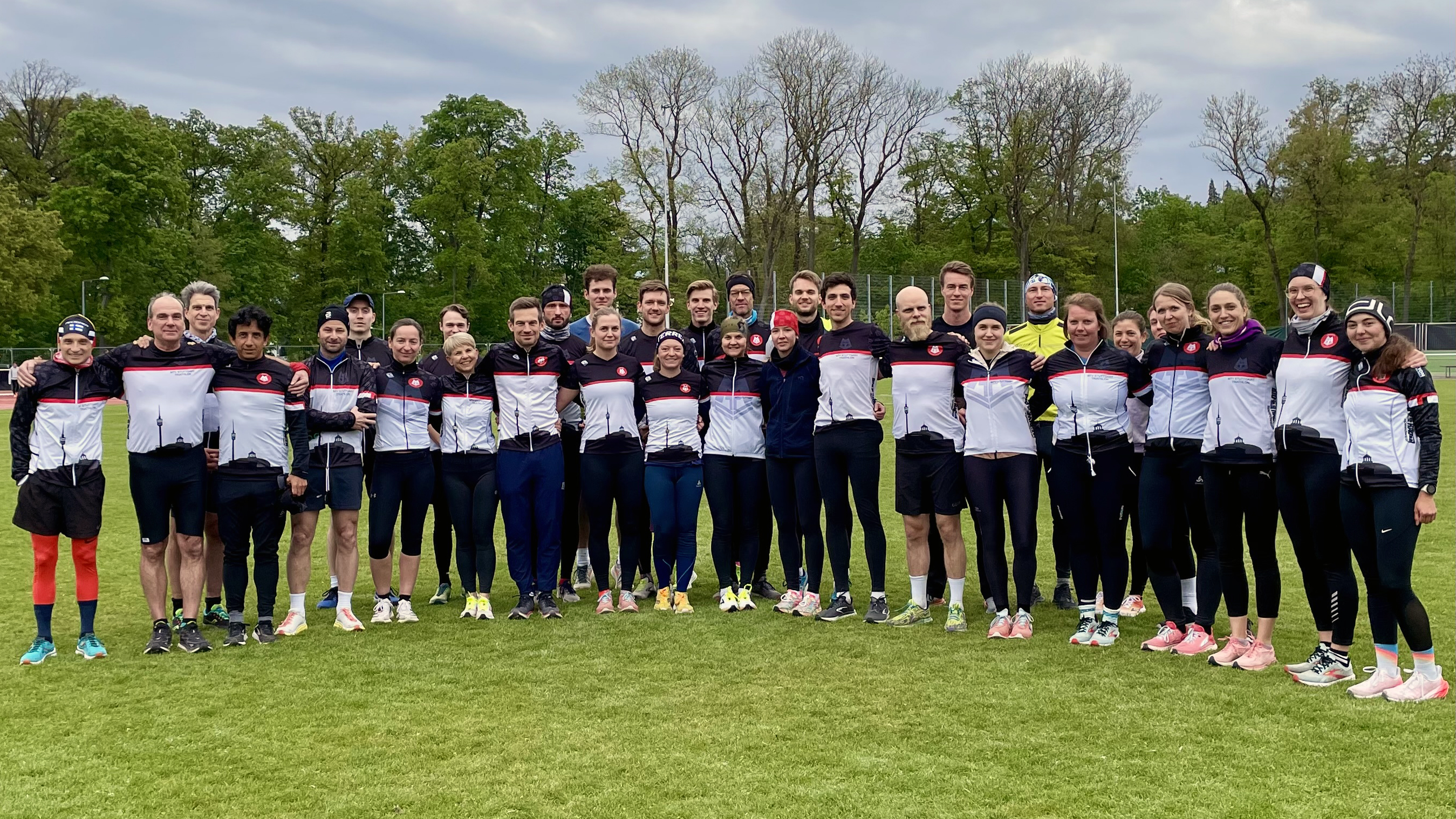 Das Bild zeigt eine Gruppe von Männern und Frauen, die in einem Halbkreis auf dem Sportplatz stehen. Bekleidet ist die Gruppen mit schwarz-weißen Trainingsjacken.